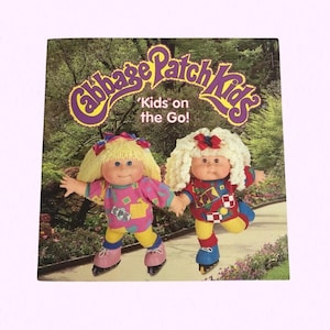 May include: A Cabbage Patch Kids book cover showing two dolls on roller skates. The dolls have blonde and white curly hair, wearing colourful outfits. The book title is "Cabbage Patch Kids" with the tagline "Kids on the Go!"