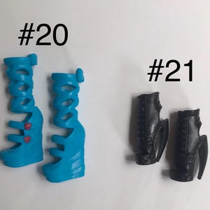 May include: Two pairs of doll shoes labeled #20 and #21. Pair #20 consists of bright blue, knee-high gladiator-style wedge sandals with two small red circular accents. Pair #21 features black, ankle-high open-toe wedge boots with lace-up detailing.