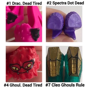 May include: Four pairs of miniature doll shoes labeled #1 Drac. Dead Tired in bright pink, #2 Spectra Dot Dead in purple, #4 Ghoul. Dead Tired in red with a black glasses graphic, and #7 Cleo Ghouls Rule in teal with gold-colored heel accents.