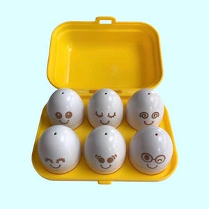 May include: A yellow plastic egg carton containing six white plastic eggs. Each egg has a different cartoon face drawn on it.
