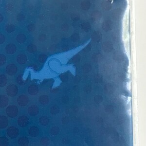Puede incluir: Una superficie rectangular azul con un patr&oacute;n de puntos azul oscuro y una silueta de dinosaurio azul claro. La palabra unique est&aacute; impresa en letras peque&ntilde;as y oscuras cerca del borde superior de la secci&oacute;n azul, con un borde verde arriba.