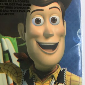 Puede incluir: Un empaque de pl&aacute;stico que presenta un primer plano del personaje Woody de Toy Story, con un sombrero de vaquero marr&oacute;n, una camisa de cuadros amarillos con un chaleco de estampado de vaca en blanco y negro y una insignia de sheriff dorada.