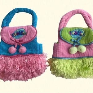 May include: Two plush toy purses. One is blue and pink with pink pom-poms and a pink fringe. The other is pink and green with green pom-poms and a green fringe. Each purse has a handle and the word "Webkinz" embroidered on the flap.