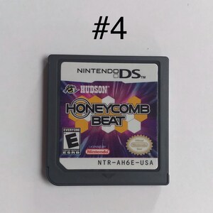 Op de afbeelding: Een Nintendo DS-gamecartridge voor "Honeycomb Beat" van Hudson. Het label van de game heeft een paars en geel honingraatontwerp met de titel van de game in vette letters. De cartridge is grijs met de ESRB-classificatie en het Nintendo-zegel.