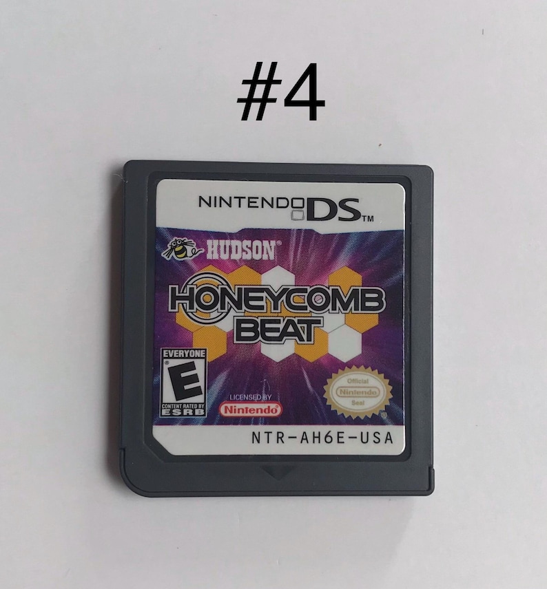 Op de afbeelding: Een Nintendo DS-gamecartridge voor "Honeycomb Beat" van Hudson. Het label van de game heeft een paars en geel honingraatontwerp met de titel van de game in vette letters. De cartridge is grijs met de ESRB-classificatie en het Nintendo-zegel.