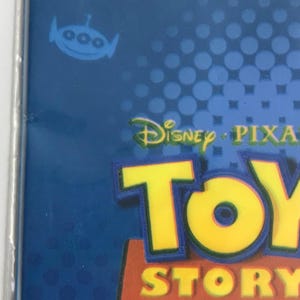 Puede incluir: Un empaque de producto azul que presenta el logotipo de Toy Story en letras amarillas y rojas. El dise&ntilde;o incluye un peque&ntilde;o gr&aacute;fico de un alien&iacute;gena azul y el texto Disney/Pixar y Poof-Slinky, Inc. impreso en la parte superior sobre un fondo azul con puntos.