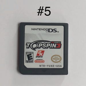 Op de afbeelding: Een Nintendo DS-gamecartridge voor Top Spin 3. De gamecartridge is donkergrijs met een wit label met de titel van de game, de ESRB-classificatie en het Nintendo-merk. De cartridge is genummerd 5.