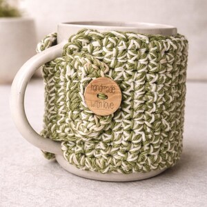 Op de afbeelding: Een keramische mok met een groen en wit gehaakte hoes. De hoes heeft een houten knoop met de tekst "handmade with love". De mok heeft een wit handvat en staat op een lichtgrijze ondergrond.