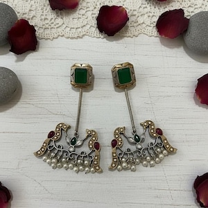May include: A pair of ornate earrings with a square green stone top and a bird-like design. The earrings feature gold and silver accents, red and green stones, and small white pearls. The background includes rose petals and a white lace.