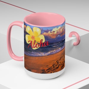 Pode incluir: Uma caneca de cerâmica rosa e branca com uma cena de praia e uma flor amarela. A caneca tem a palavra "Aloha" escrita em vermelho.