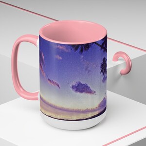 Pode incluir: Caneca de cerâmica branca com alça e interior rosa. A caneca apresenta uma imagem panorâmica de um pôr do sol sobre o oceano com palmeiras e nuvens. O céu é uma mistura de tons de azul, roxo e rosa.