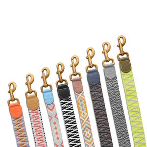 Op de afbeelding: Een verzameling kleurrijke hondenriembanden met goudkleurige metalen sluitingen. De banden hebben verschillende patronen en kleuren, waaronder oranje, roze, zwart en grijs. Elke band is schuin geplaatst, wat de verschillende ontwerpen en texturen benadrukt.