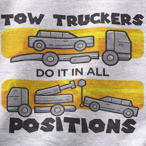 Może przedstawiać: Szara koszulka z żółtym tłem i czarnym nadrukiem przedstawiającym holownik. Nadruk głosi "Tow Truckers Do It In All Positions".