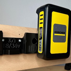 Könnte beinhalten: Eine schwarze Wandhalterung aus Kunststoff mit dem Text "Kärcher 18/36V" und ein schwarzer und gelber Akku mit dem Text "Kärcher Battery Power 18V/3 2.5 Ah", der an einer Holzoberfläche befestigt ist.