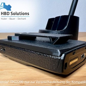 Könnte beinhalten: Ein schwarzes elektronisches Gerät mit strukturierter Oberfläche, mit einem Ladeanschluss und einem kleinen, rechteckigen Bildschirm. Das Gerät befindet sich auf einer Holzoberfläche, mit einem schwarzen Ständer und einem Logo im Hintergrund. Der Text "HBD Solutions" ist sichtbar.