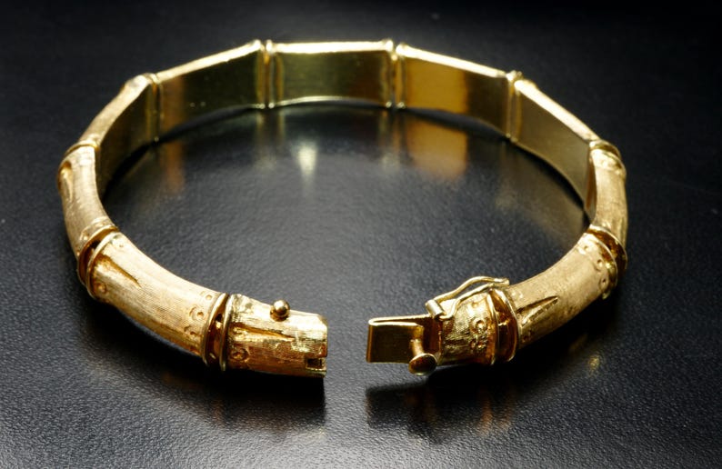 May include: A gold bamboo-style bangle bracelet with a hinged clasp. The bracelet is made of polished gold and has a textured finish.