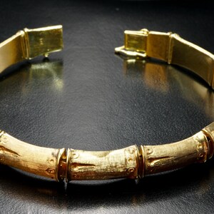 May include: A gold-toned bangle bracelet with a bamboo-inspired design. The bracelet is made of smooth, polished metal and features a series of raised, rounded segments that resemble bamboo stalks.