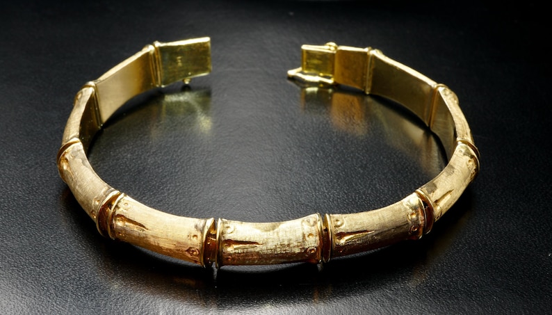 May include: A gold-toned bangle bracelet with a bamboo-inspired design. The bracelet is made of smooth, polished metal and features a series of raised, rounded segments that resemble bamboo stalks.