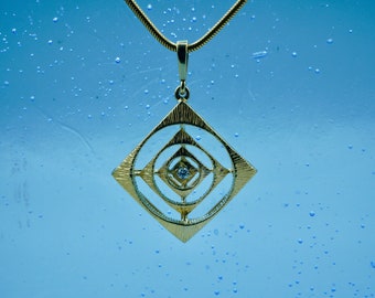 May include: A gold pendant necklace with a geometric design featuring a small diamond in the center. The pendant is hanging against a blue background with small white dots.