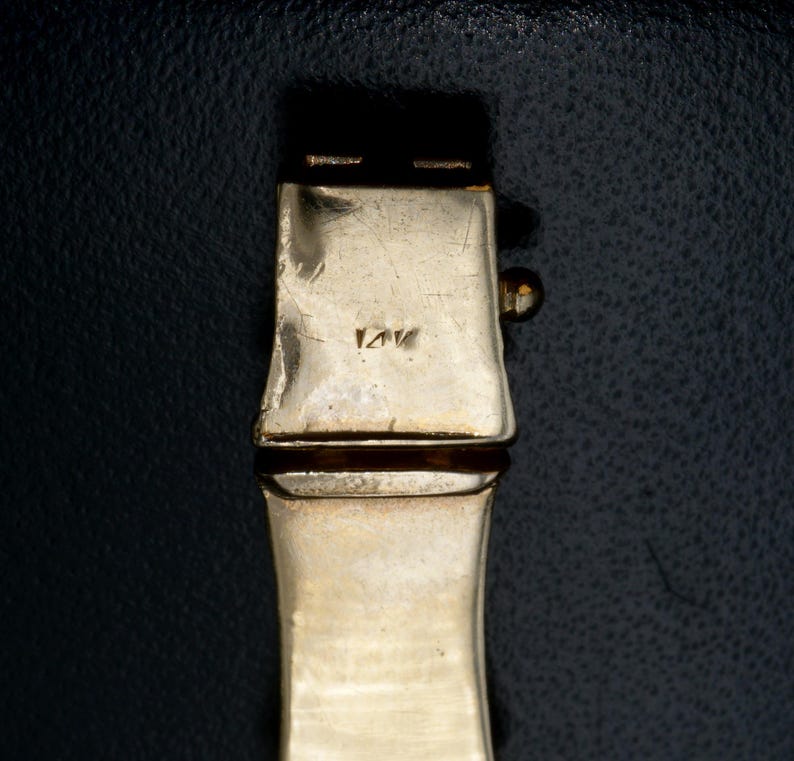 May include: Close-up of a gold-toned metal bracelet clasp with the inscription "14K" on the back.