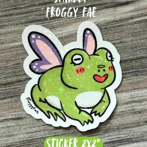 May include: A sparkly green frog sticker with purple and pink fairy wings. The sticker has the text "Sparkly Froggy Fae" and "Sticker 2x2". The frog has a smiling red mouth and rosy cheeks.