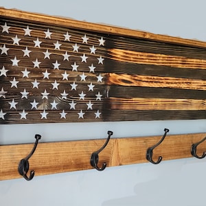 May include: A wooden coat rack with a distressed American flag design. The rack has four black metal hooks and a brown stained finish.