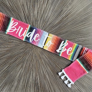 May include: A pink and multi-coloured fabric sash with white fringe and the text "Bride to Be" in white lettering.