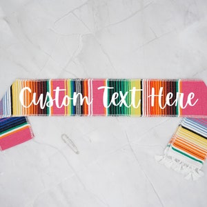 May include: A colorful Mexican-style serape sash with white text that reads "Custom Text Here".