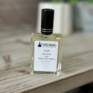 May include: A clear glass perfume bottle with a black cap. The label reads "Lush Inspired by Lush Twenty Nine High St Eau de Parfum 30ml | 1oz". The bottle is on a wooden surface.