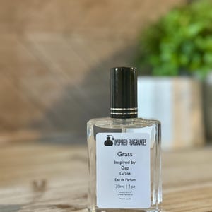 May include: A clear glass perfume bottle with a black cap and a white label. The label reads "Grass Inspired by Gap Grass Eau de Parfum 30ml | 1oz". The bottle is on a wooden surface.