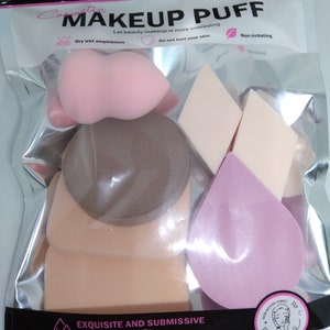 May include: A clear plastic bag containing various makeup puffs in shades of pink, beige, and brown. The bag has text that reads "MAKEUP PUFF" and "EXQUISITE AND SUBMISSIVE BEAUTIFUL & QUALITY". The puffs are in different shapes, including teardrops and rounded shapes.