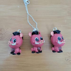 Puede incluir: Tres dijes de lechón rosa y negro con ojos grandes y colmillos. Los dijes están unidos a cadenas plateadas. Los dijes están sobre una superficie de madera.