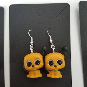 May include: A pair of dangle earrings featuring yellow cartoon puppies with black ears and large, sparkly eyes. The puppies are wearing green collars. The earrings are made of silver.