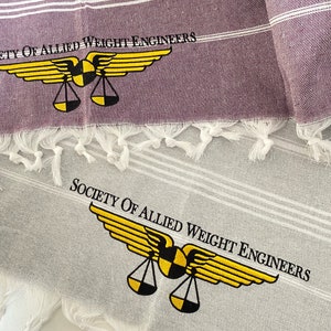 May include: Two white towels with gray stripes and fringed edges. The top towel is purple and has the text "Society of Allied Weight Engineers" and a yellow winged scale logo. The bottom towel has the same logo and text in black.