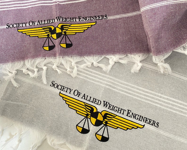 May include: Two white towels with gray stripes and fringed edges. The top towel is purple and has the text "Society of Allied Weight Engineers" and a yellow winged scale logo. The bottom towel has the same logo and text in black.