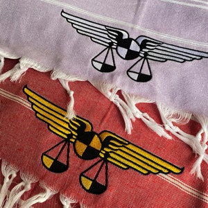 May include: Two embroidered towels with a design of a pair of wings and scales. The top towel is light purple with a white design. The bottom towel is red with a yellow design.