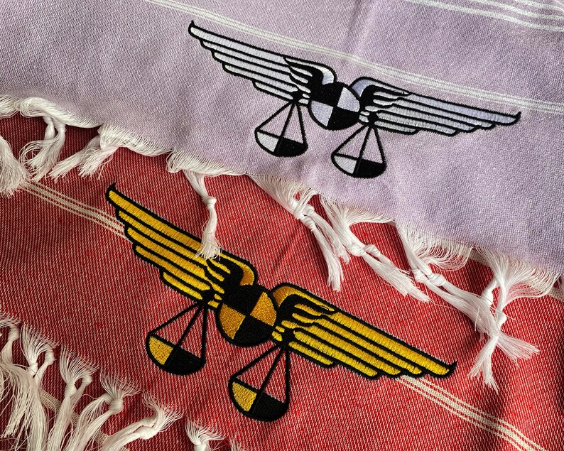 May include: Two embroidered towels with a design of a pair of wings and scales. The top towel is light purple with a white design. The bottom towel is red with a yellow design.