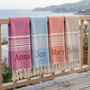 May include: Four rectangular cotton towels with fringe trim, each featuring a different colour: magenta, light blue, peach, and beige. Each towel displays a white horizontal striped pattern and is embroidered with a name: Anna, Jen, Mary, or Debbie.