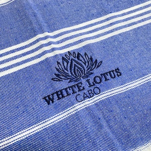 May include: A blue and white striped beach towel with the embroidered text "White Lotus Cabo" and a stylized lotus flower design.