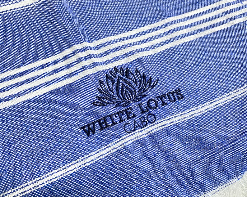 May include: A blue and white striped beach towel with the embroidered text "White Lotus Cabo" and a stylized lotus flower design.