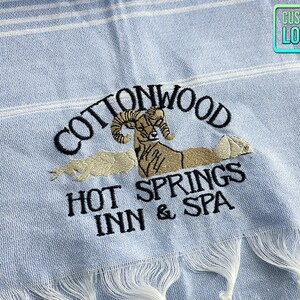 May include: A light blue beach towel with white stripes and an embroidered logo that says "Cottonwood Hot Springs Inn & Spa" with a ram's head in the center.