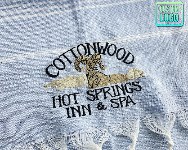 May include: A light blue beach towel with white stripes and an embroidered logo that says "Cottonwood Hot Springs Inn & Spa" with a ram's head in the center.