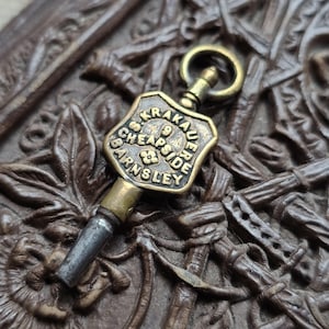 May include: A vintage brass watch key with a shield-shaped head. The head is engraved with the words "S. KRAKAUE, CHEAPSIDE, BARNSLEY" and the number "9". The key has a small loop at the top and a silver-colored tip.