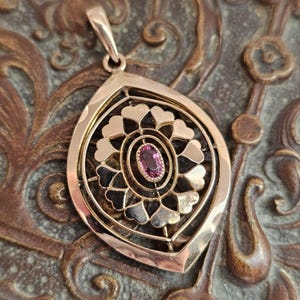 May include: A gold pendant featuring a central oval-cut pink gemstone surrounded by a decorative openwork frame of heart-shaped motifs. The pendant has a pointed, teardrop-shaped outer border and a small bail at the top for attachment to a chain.