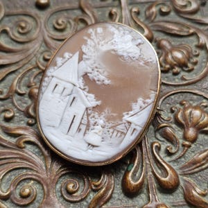 May include: An oval-shaped cameo brooch featuring a white carved relief of a small village scene with a church and a figure on a light brown background, encased in a simple gold-toned metal frame with a smooth, polished edge finish.