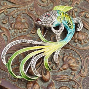 May include: A detailed, silver-toned fish brooch with marcasite accents and colorful enamel. The fish features a red mouth, a marcasite-studded head, and a body in shades of blue and green. The long, flowing tail is a mix of green and yellow.