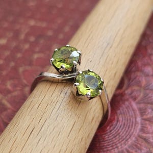 May include: A bypass ring crafted from silver-toned metal featuring two round-cut, faceted lime green peridot gemstones. Each stone is secured in a six-prong setting, with the band curving to hold the stones in a staggered, open arrangement.