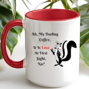 Puede incluir: Una taza de cerámica blanca con interior y asa de color rojo brillante. El frente presenta una ilustración de una mofeta en blanco y negro junto al texto negro: Ah, My Darling Coffee, It Is Love At First Sight, No? La palabra Love es roja.