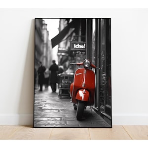 May include: A red Vespa scooter parked on a cobblestone street in a black and white city setting. The scooter is the only color in the image.