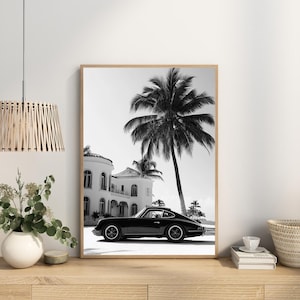May include: Black and white photograph of a classic car parked in front of a white house with palm trees. The car is a vintage Porsche 911.